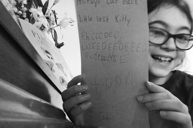 girl with glasses holds a piece of paper that says "Little Lost Kitty" "R.H. CCCDEC L.H. CEEDEEDEEEC"... it's a piano composition!