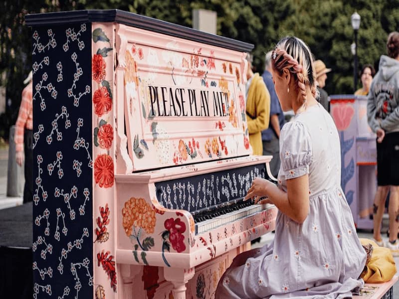 Person in white daisy dress plays upright piano painted pink and black and covered in painted flowers